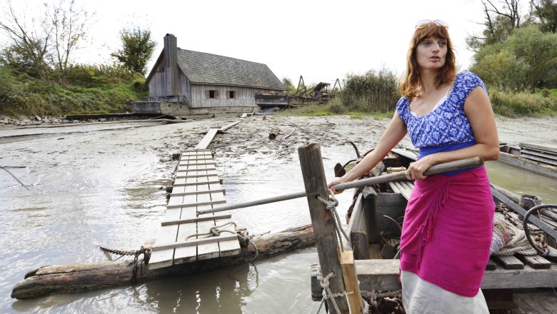 Frau in traditioneller Kleidung auf einem Boot vor einem Holzhaus am Flussufer., © Donau Niederösterreich, Steve Haider